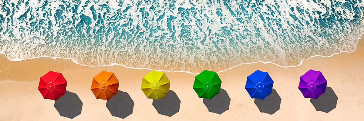 Aerial photograph of colorful umbrellas in a rainbow pattern on a beach on Fire Island, NY with ocean waves by Evan Reinheimer.