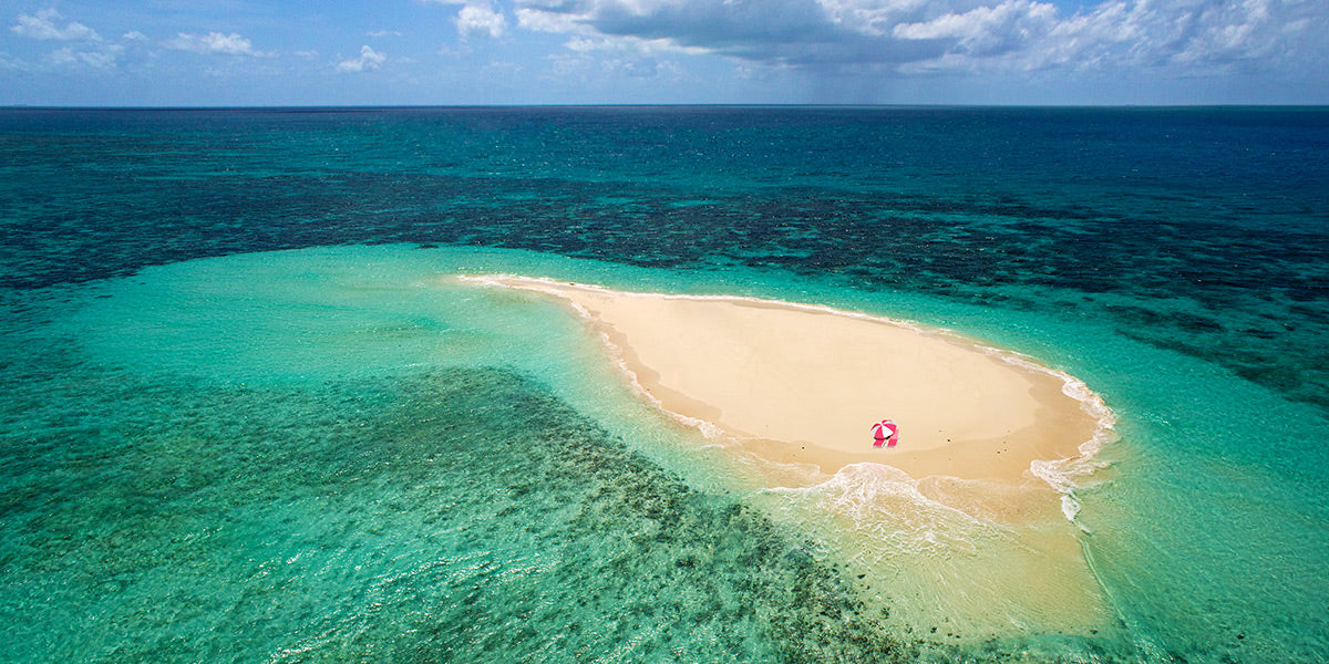 Aerial photograph of island with a pink umbrella surrounded by clear turquoise water and blue sky by Evan Reinheimer.