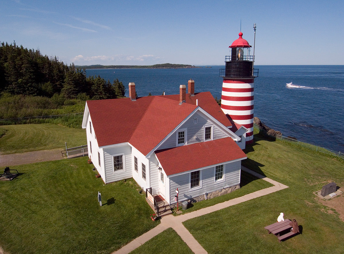 Aerial photo of West Quoddy Lighthouse with a red roof and white stripes on a grassy area near water in Lubec, Maine by Evan Reinheimer.