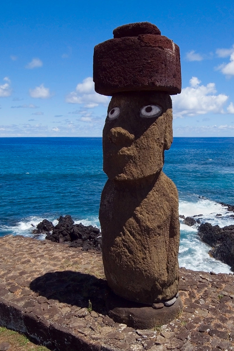 Aerial photograph of a Moai stone statue with a hat against a coastal background on Easter Island by Evan Reinheimer.